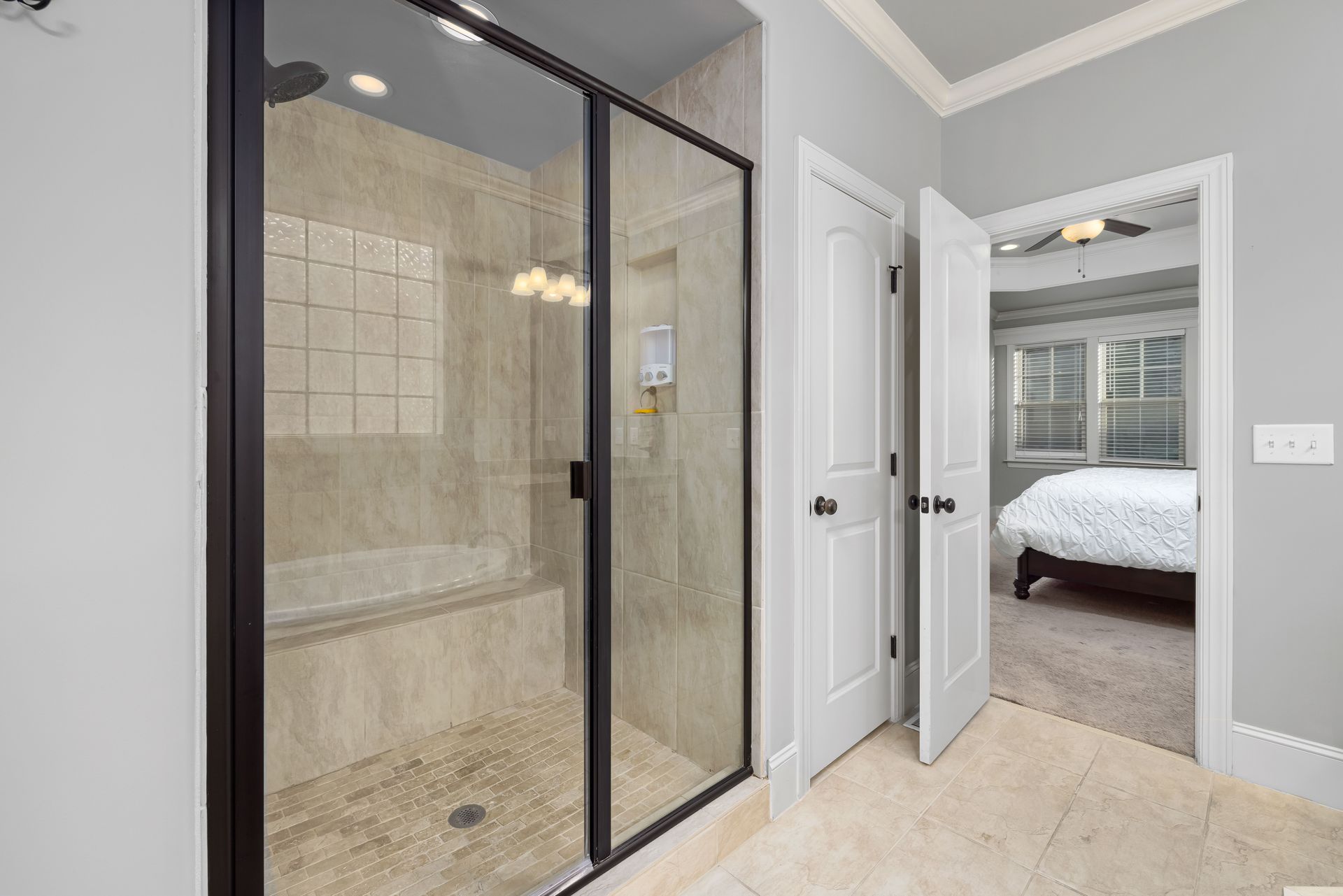 An upscale bathroom featuring a sleek glass shower door, leading to a cozy bedroom with natural lighting.